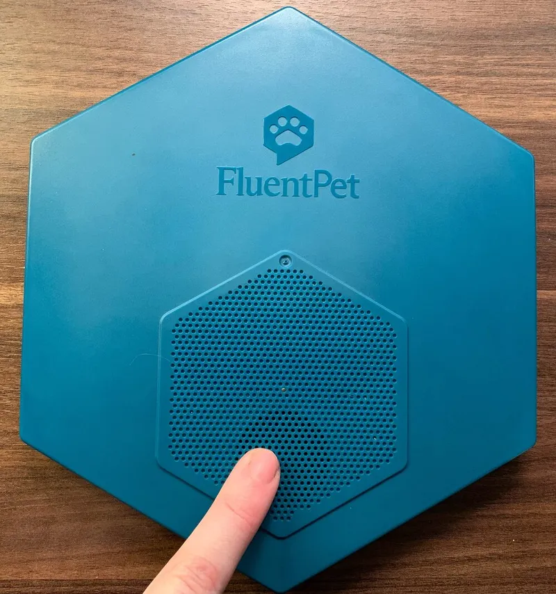 The top of a Connect Base, with a finger pointing at the hexagonal speaker grill below the FluentPet logo.