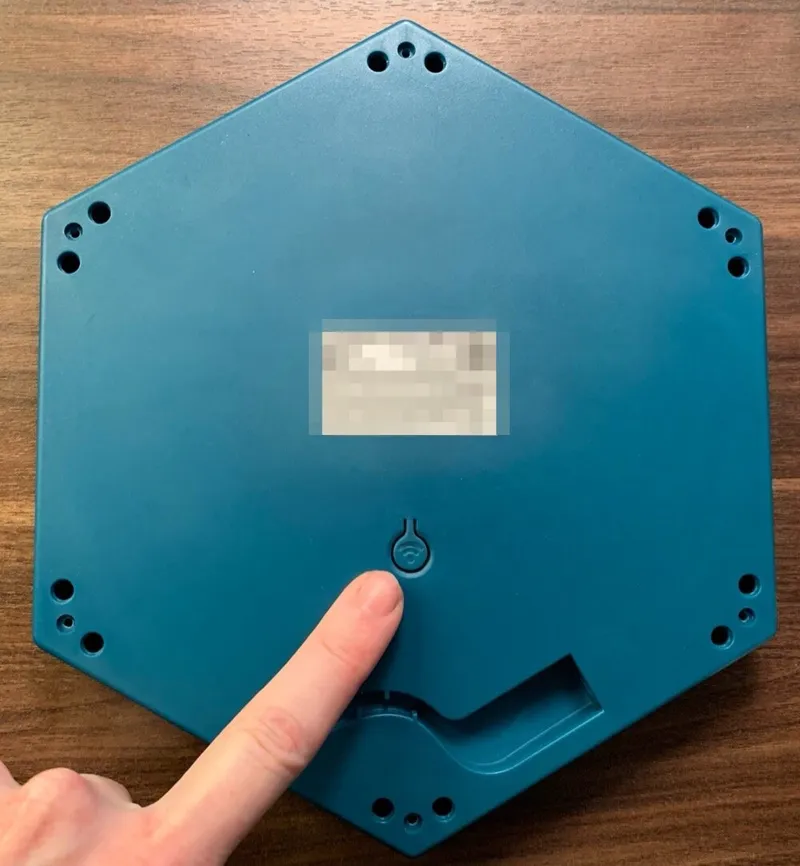 The bottom of a Connect Base, with a finger pointing at the Base Button, marked with a Wi-Fi symbol, below the product label. The label is blurred.